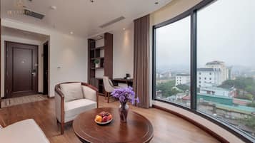 Quarto de hotel moderno com uma janela grande, uma mesa de madeira redonda com frutas e flores, um sofá e uma escrivaninha.