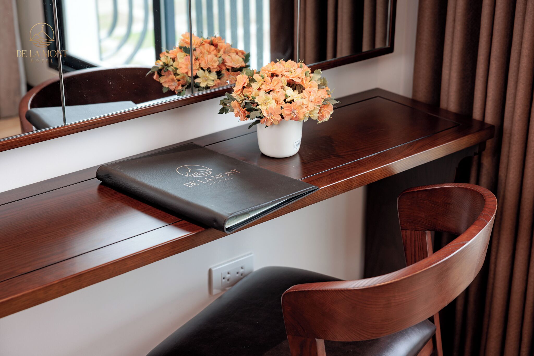 A wooden desk with a white vase of flowers, a black folder, and a chair.