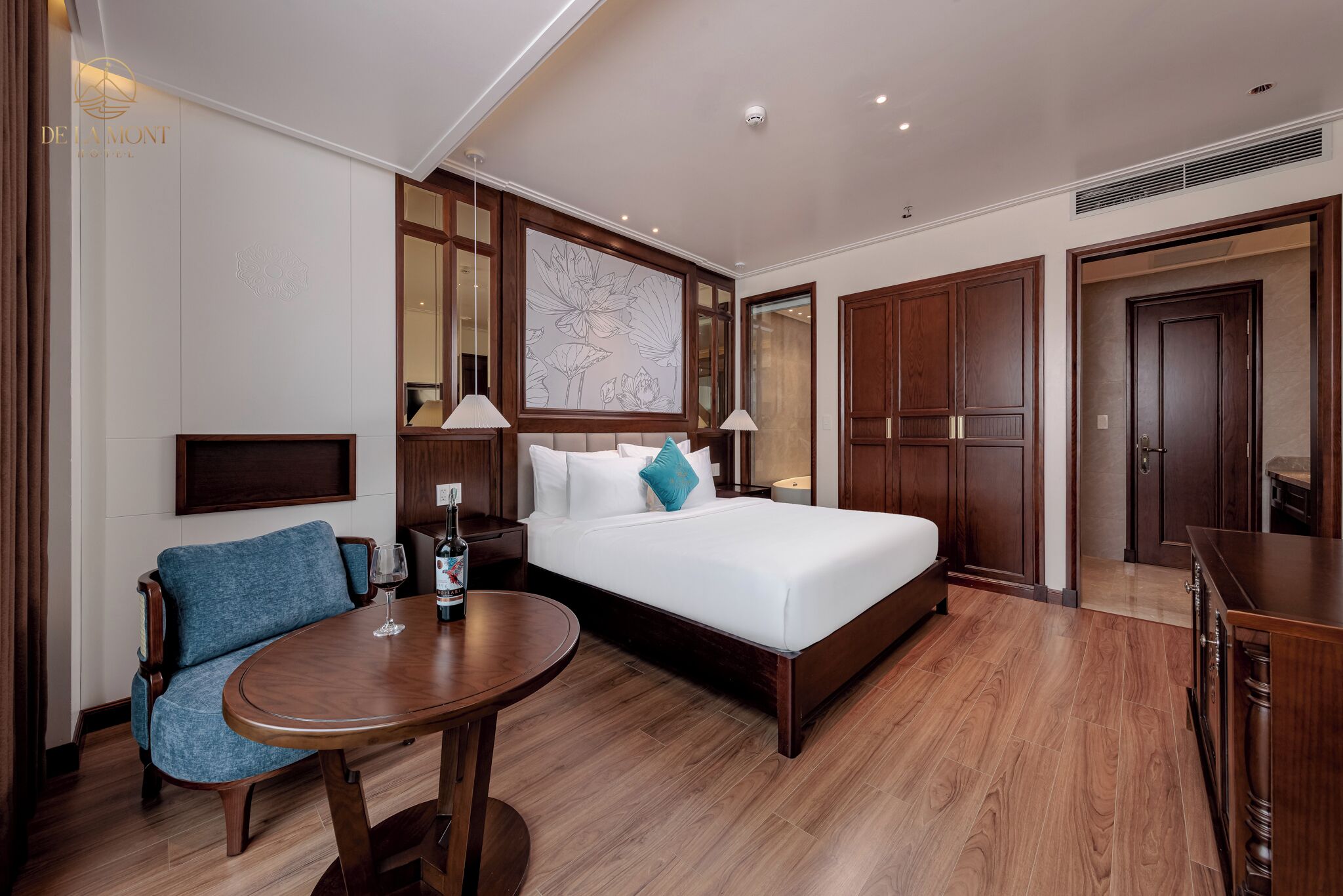 Una camera d'albergo con un letto grande, un comodino in legno, una poltrona blu, un tavolino rotondo in legno e un'ampia opera d'arte incorniciata appesa al muro.