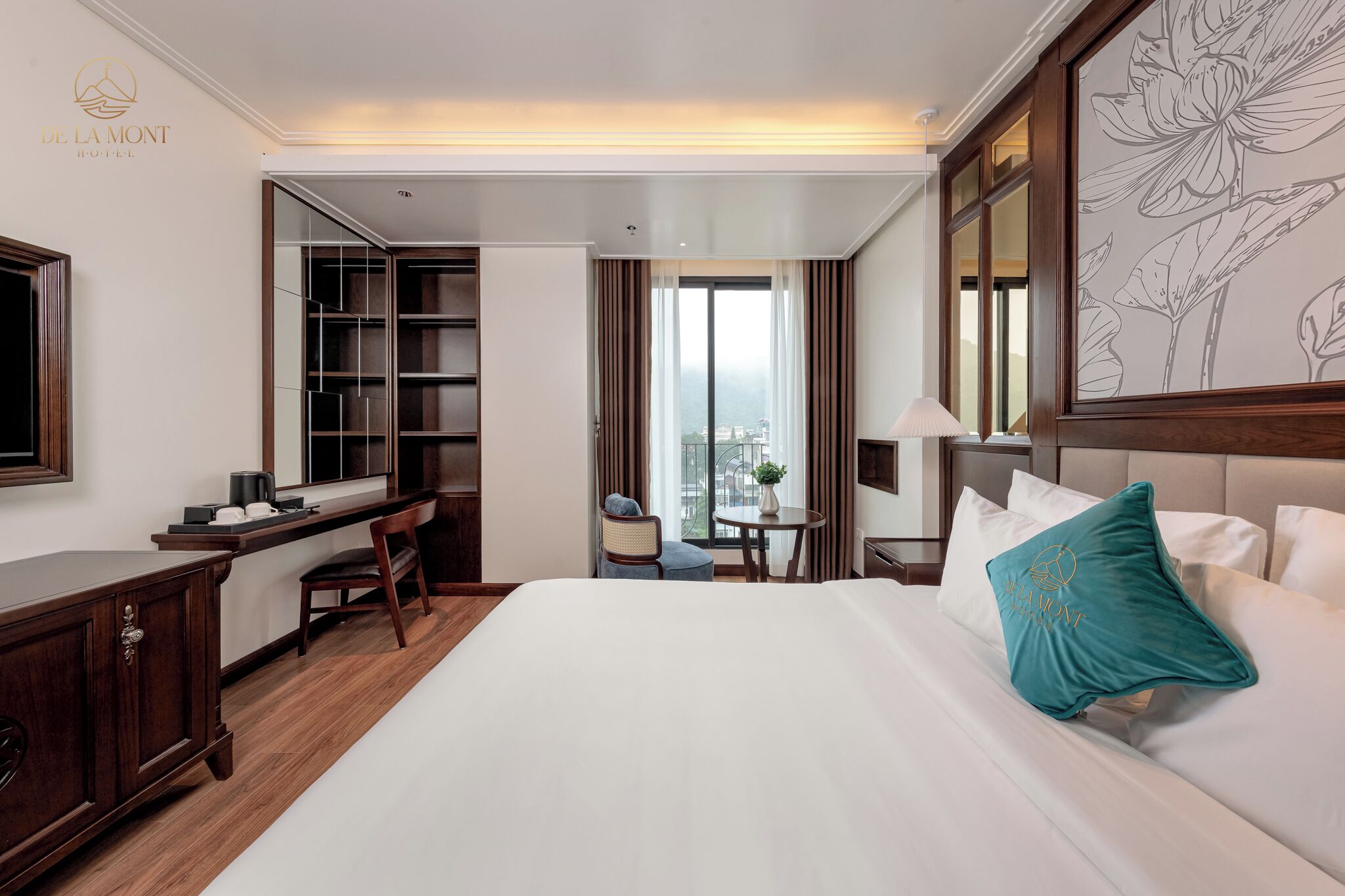 A hotel room with a large bed, a desk, a chair, a small table with a lamp, and a view of the city through large windows.