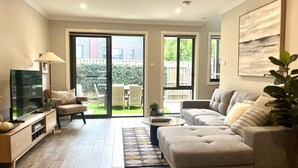 A modern living room with a sofa, a coffee table, and a TV. There are sliding glass doors leading to a patio with outdoor furniture. - Stylish Townhouse With Private Courtyard (Canberra)