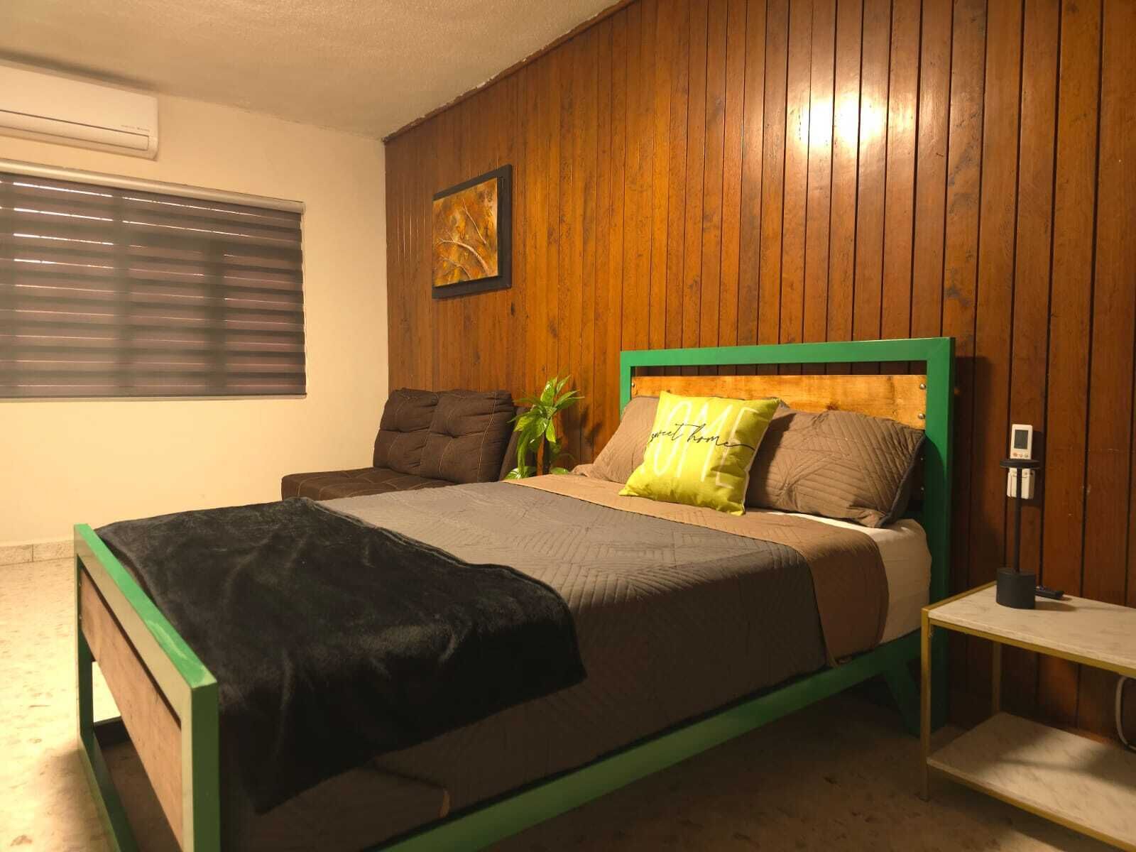 Een slaapkamer met een wand van houten panelen, een bed met groen en bruin beddengoed, een nachtkastje en een raam met zonwering.