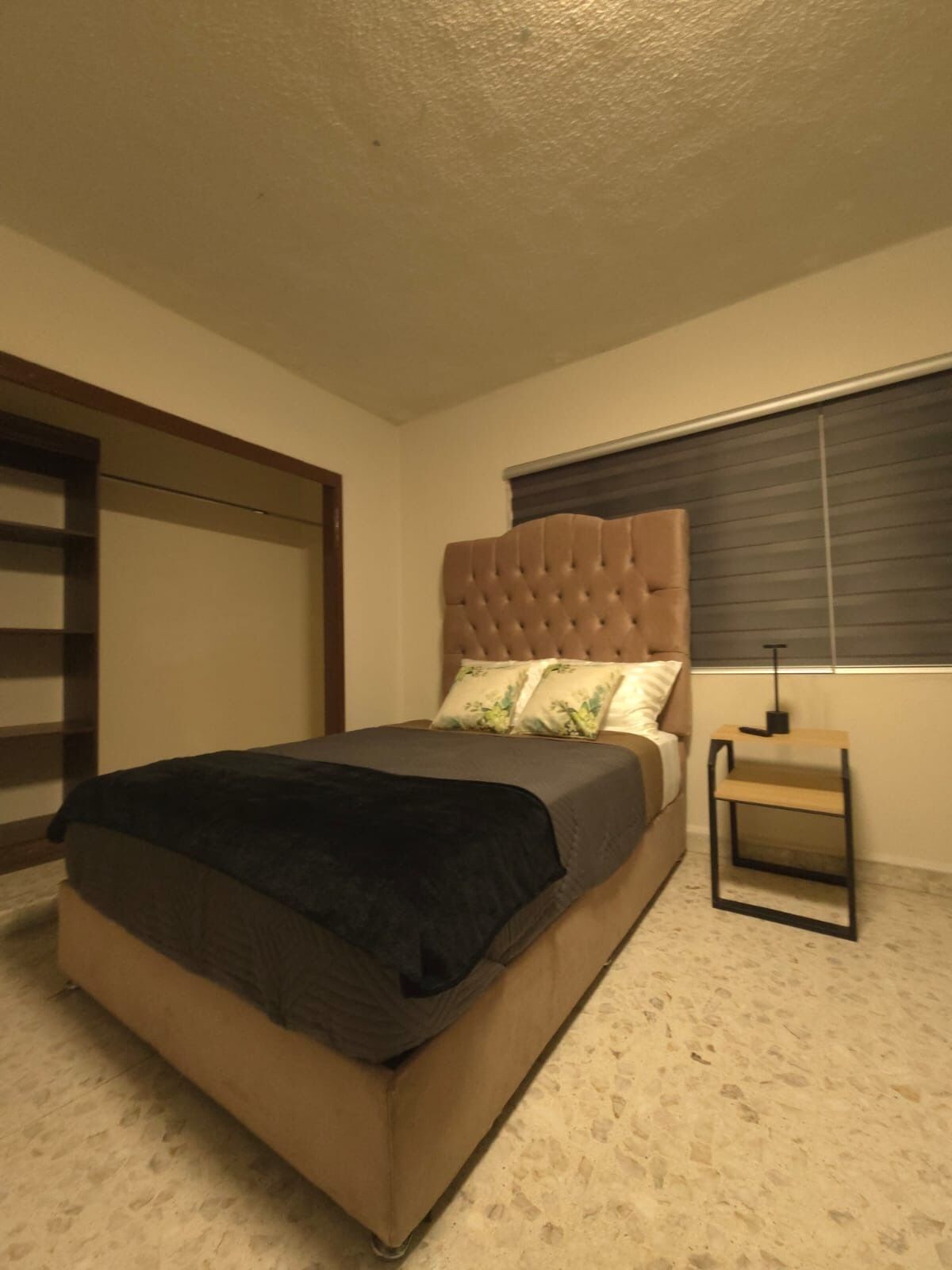 Un dormitorio con una cama, una mesita de noche, una ventana con persianas y un cabecero de madera.