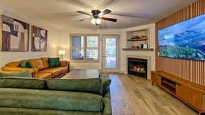 A living room with a large flat-screen TV, a ceiling fan, a fireplace, and a green couch. - Downtown Helen - 1st Floor | Free Parking (Helen)