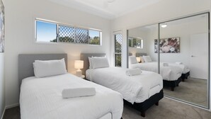 Habitación de hotel con dos camas, ventilador de techo, espejo grande y ventana con vistas a árboles.