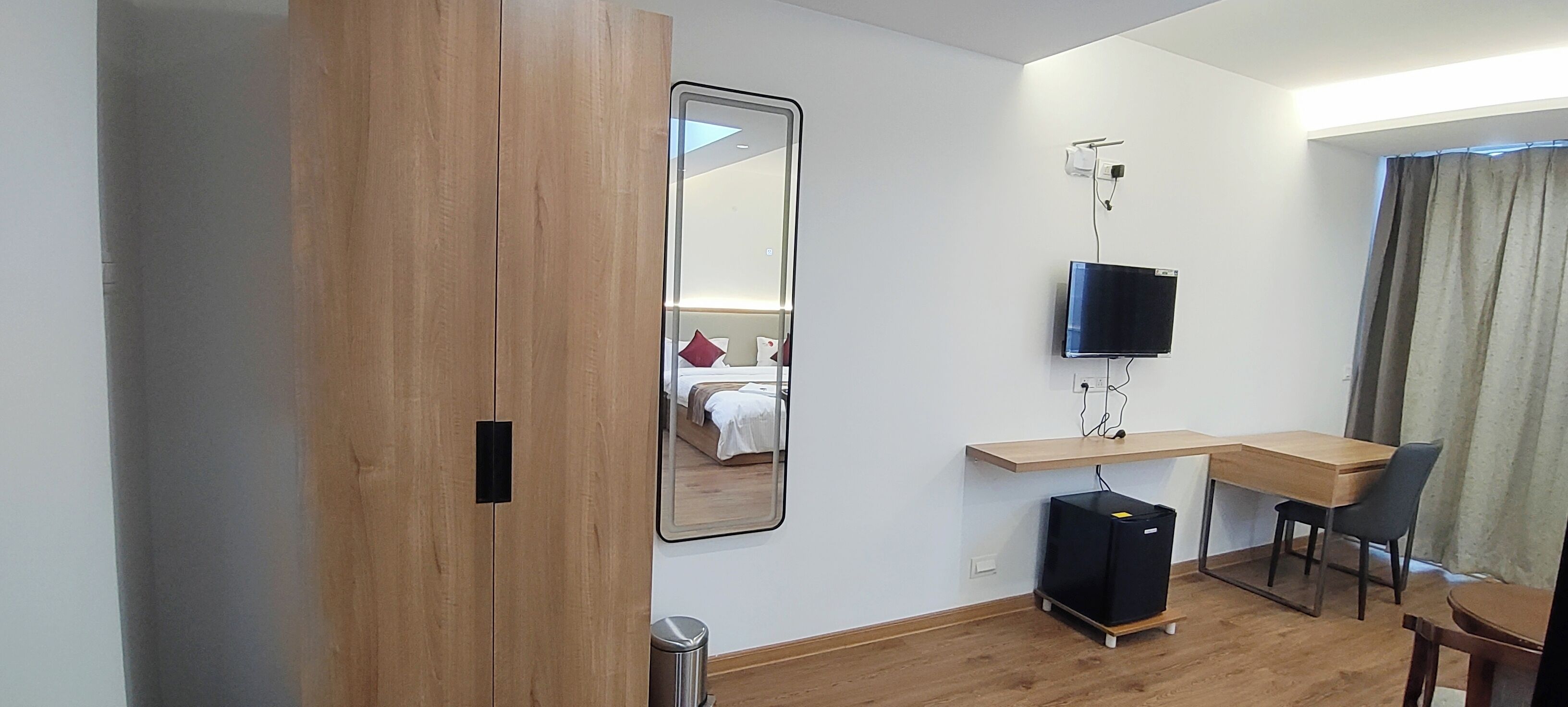 Ein Hotelzimmer mit einem Holzschrank, einem Schreibtisch mit Stuhl, einem an der Wand befestigten Fernseher und einem Bett mit rotem Kissen.