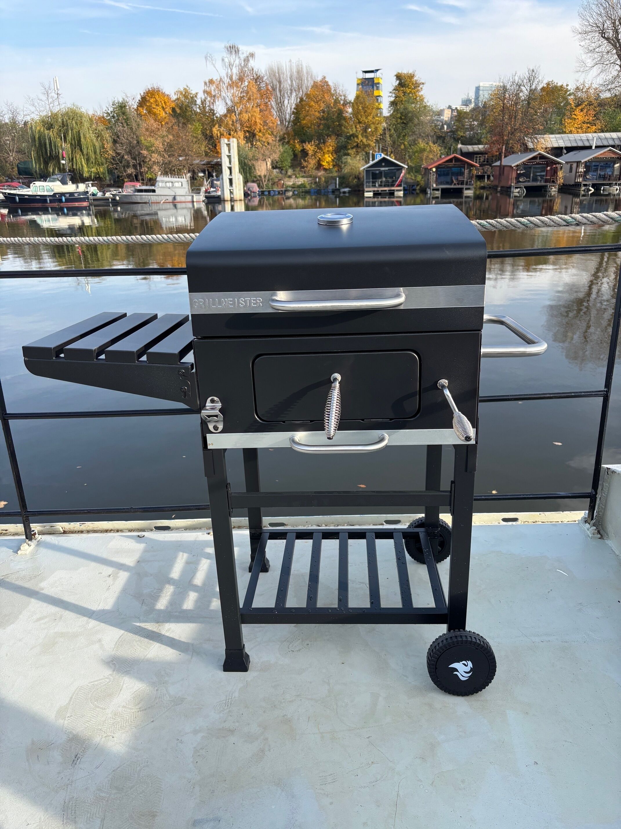 A portable grill with a handle and wheels, placed on a concrete surface with a view of a body of water and buildings in the background.