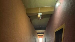 A long, narrow hallway with a tiled floor and walls painted in a reddish-brown color. There is a ceiling fan and a light fixture visible. - Hotel Raghukul (Shri Madhopur)