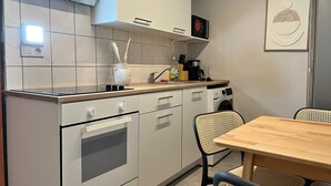 A modern kitchen with white cabinets, a built-in oven, a coffee maker, and a dining area with a wooden table and woven chairs. - Lovely Studio for 4 with Garden (Vienna)