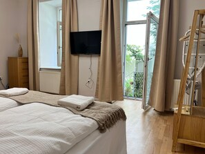 Un dormitorio con cama, televisor montado en la pared, cómoda de madera y ventana con cortinas.