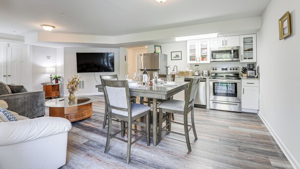 A modern kitchen with stainless steel appliances, a dining area with a glass-top table, and a living room with a flat-screen TV.