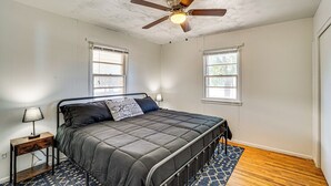 Un dormitorio con una cama, ventilador de techo, dos ventanas, una mesita de noche y una alfombra estampada.