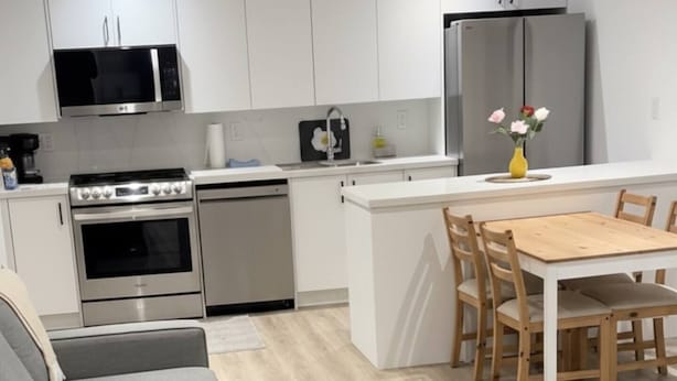 Una cocina moderna con armarios blancos, horno empotrado, microondas y refrigerador. Hay un comedor con una mesa y sillas de madera, y un sofá gris.