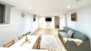 Una sala moderna con un sofá, una mesa de comedor con sillas, un televisor montado en la pared y un reloj en la pared.