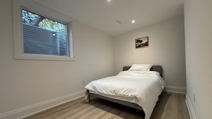 Un dormitorio con una cama, una ventana con persianas, una lámpara en el techo y un cuadro pequeño en la pared.