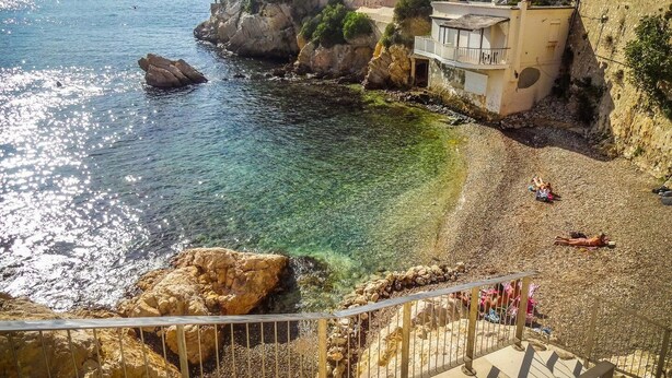 Vista costeira com águas cristalinas, praia de pedras e edifícios no topo de um penhasco.