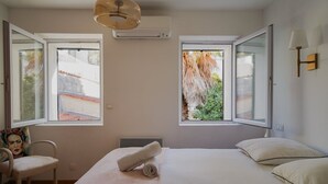 Quarto com cama, duas janelas, uma cadeira, luminária e ar condicionado.