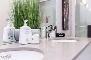 Een badkamer met een wit wastafel, een spiegel en toiletartikelen, waaronder een fles bodylotion, wattorondjes en een make-up tas.