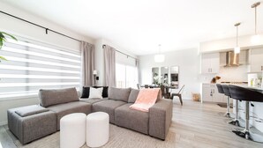 A modern living room with a sectional sofa, ottomans, a dining area with a table and chairs, and a kitchen with bar stools. - Stunning 4BD Home | Gym | Yard | AC | Games Room! (Edmonton)