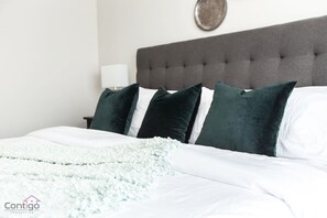 Een net opgemaakt bed met witte lakens, een lichtgroene deken, een donkergrijs gestuikt hoofdbord en drie donkergroene kussens.