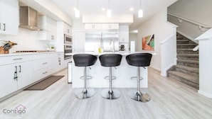 A modern kitchen with white cabinetry, a central island with bar stools, and a staircase leading to a loft area. - Stunning 4BD Home | Gym | Yard | AC | Games Room! (Edmonton)