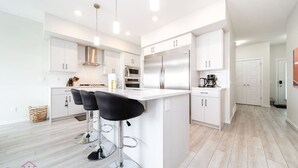 A modern kitchen with white cabinetry, stainless steel appliances, and black bar stools. - Stunning 4BD Home | Gym | Yard | AC | Games Room! (Edmonton)