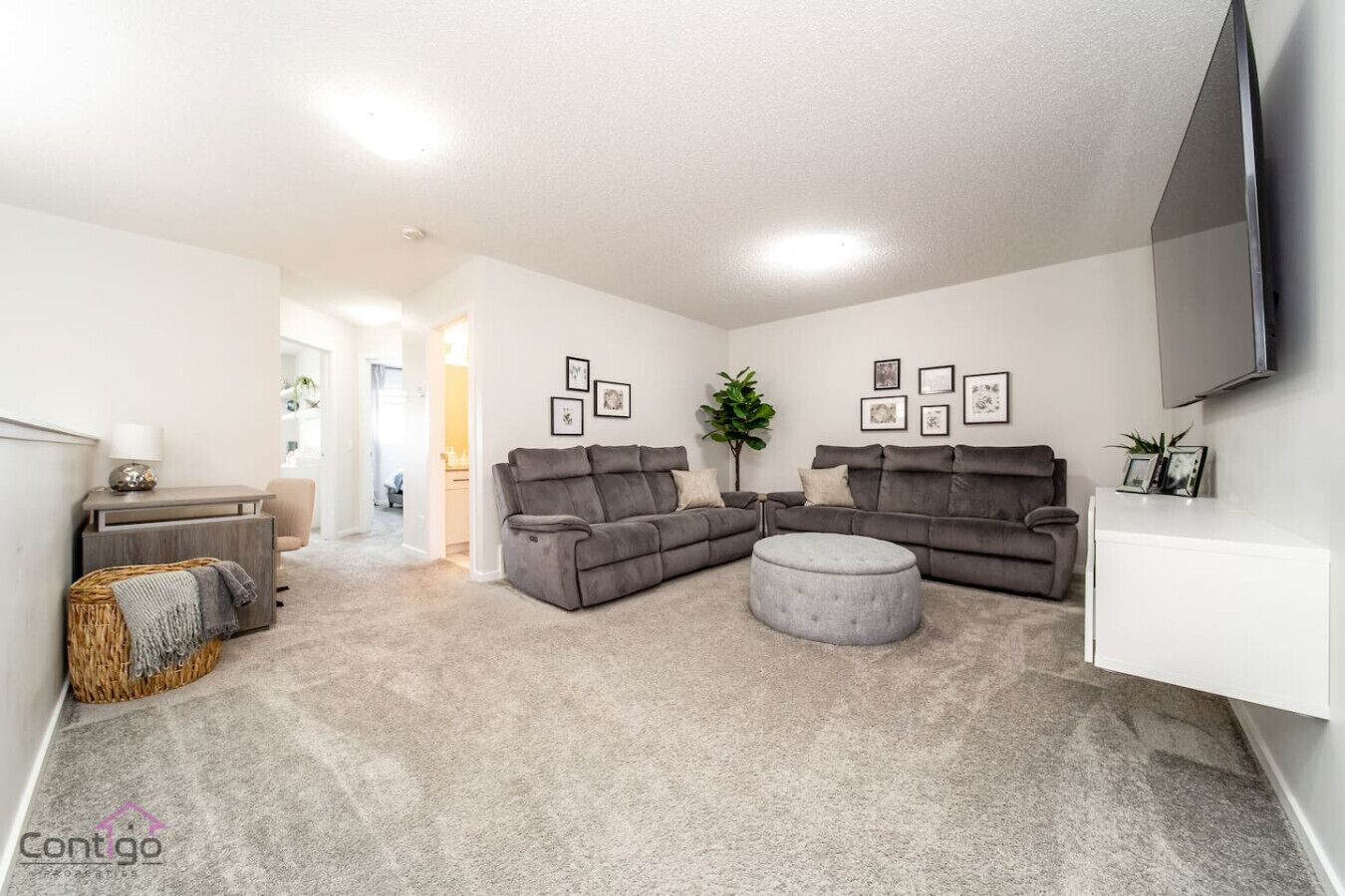 A modern living room with a gray sectional sofa, a round ottoman, and a flat-screen TV mounted on the wall.