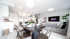 A modern kitchen with white cabinets, a dining table set for four, and a grey sofa in the living area. - Stunning 4BD Home | Gym | Yard | AC | Games Room! (Edmonton)