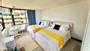 A hotel room with two beds, a small desk, a chair, and a view of the city. - Cozy Ocean-view studio in the heart of Waikiki (Honolulu)