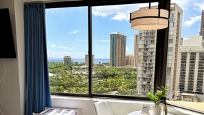 A hotel room with a bed, a small table with chairs, a view of the cityscape, and a window with curtains. - Cozy Ocean-view studio in the heart of Waikiki (Honolulu)
