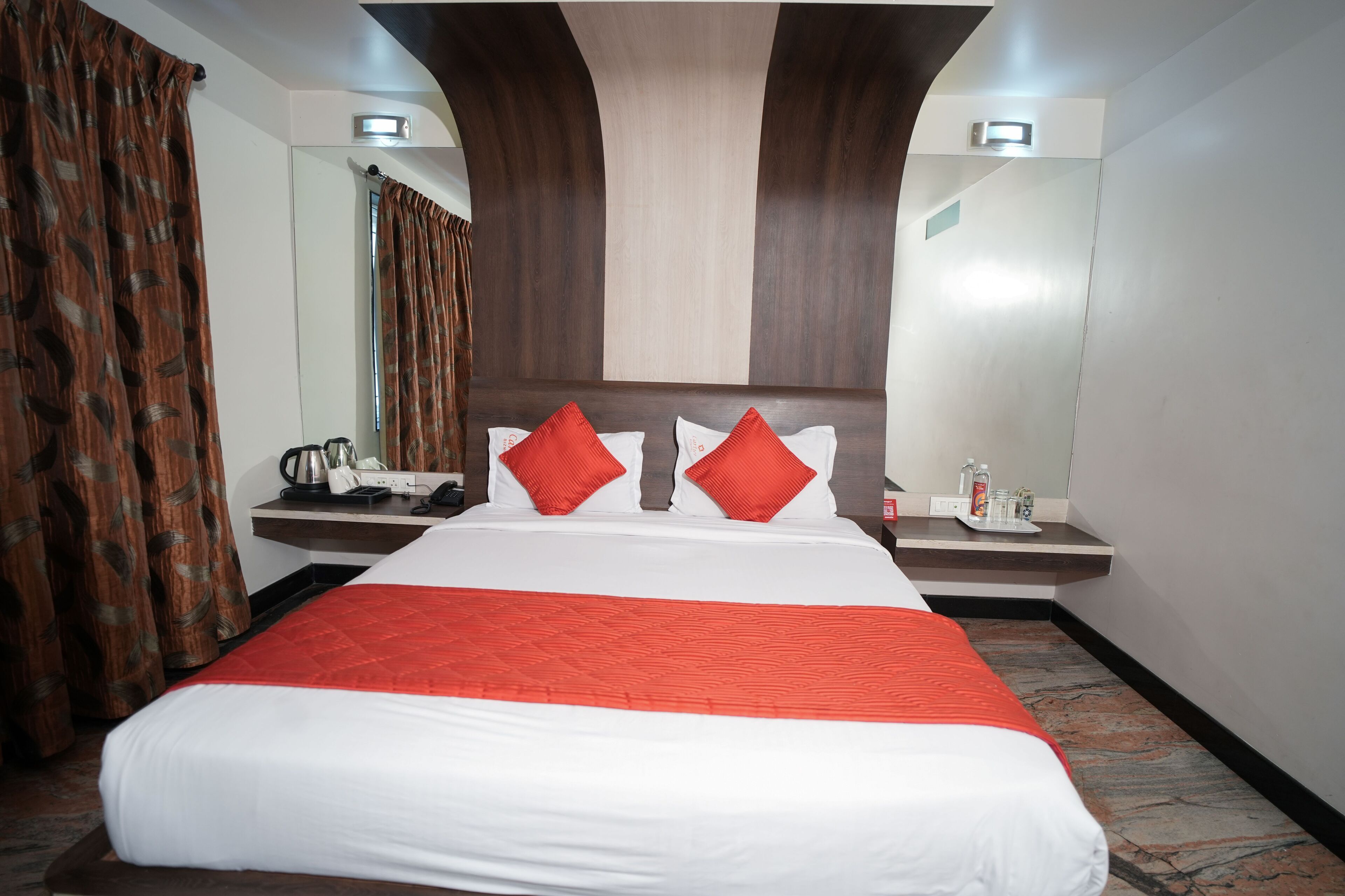 A hotel room with a large bed, two red pillows, a white bedspread, a nightstand with a lamp, a mirror, and a window with curtains.