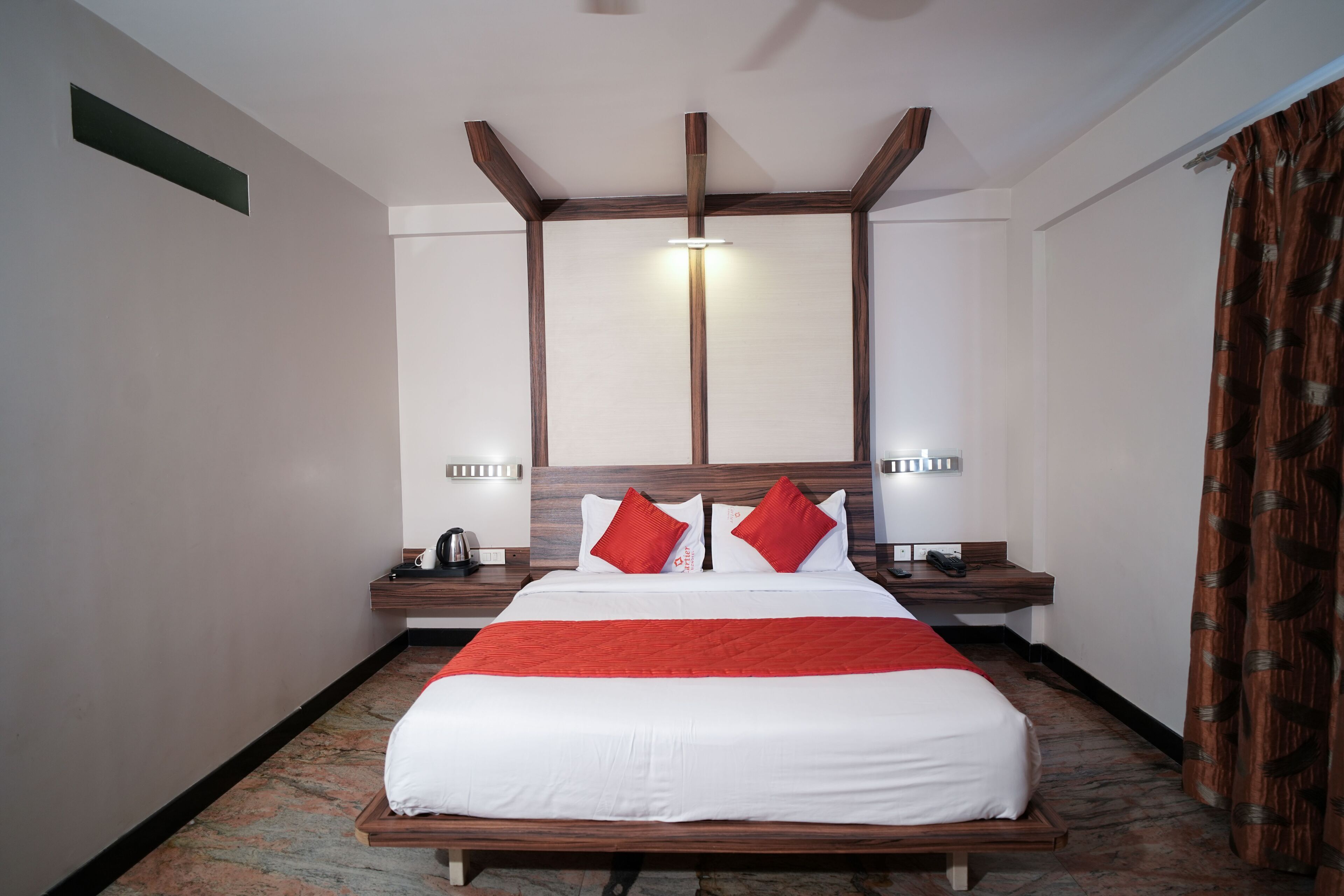 A hotel room with a large bed, wooden headboard, two bedside tables, a nightstand, and a red throw blanket.