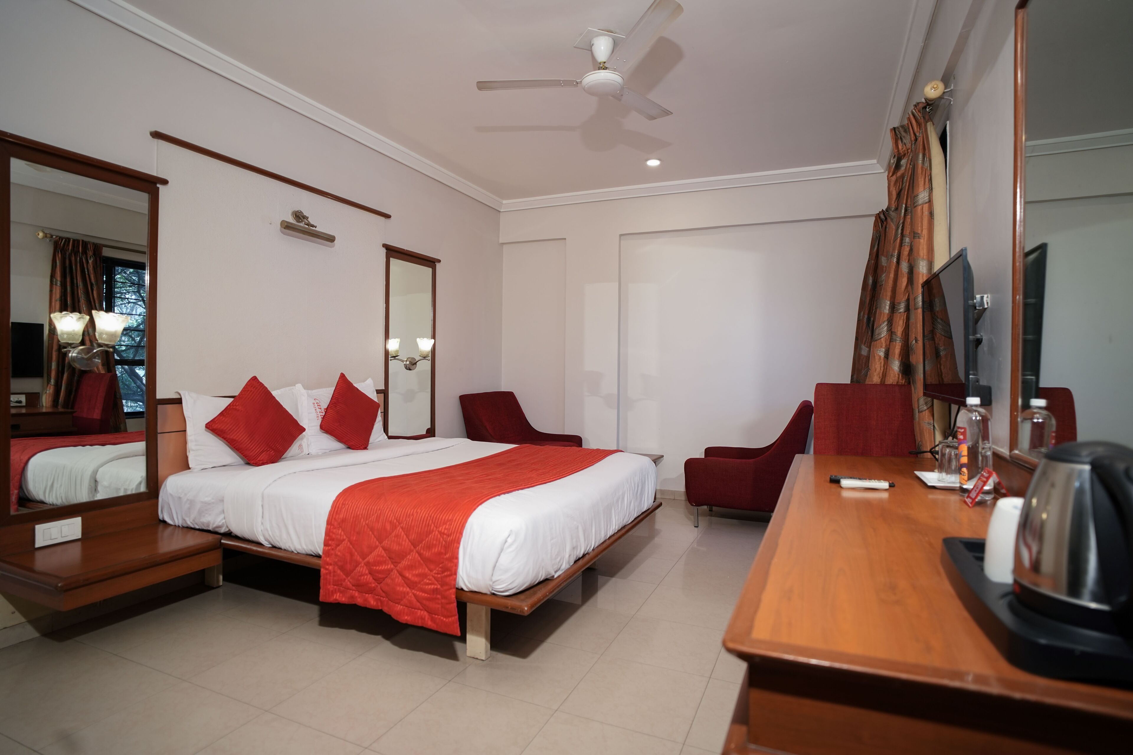 A hotel room with a large bed, two red chairs, a wooden desk, a ceiling fan, and a television.