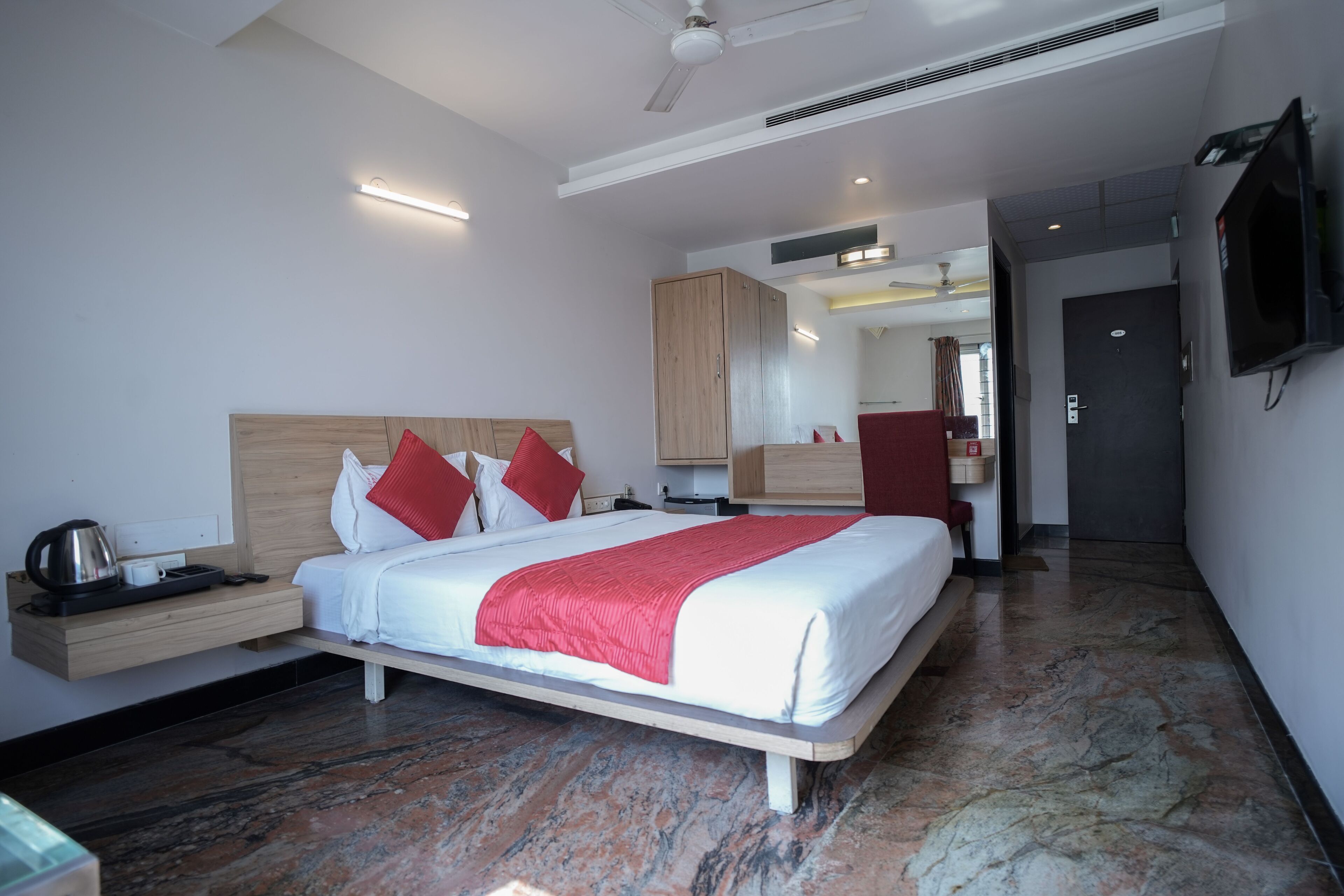 A modern hotel room with a large bed, wooden headboard, and a red throw blanket. There is a bedside table with a kettle and a tray, and a flat-screen TV mounted on the wall.
