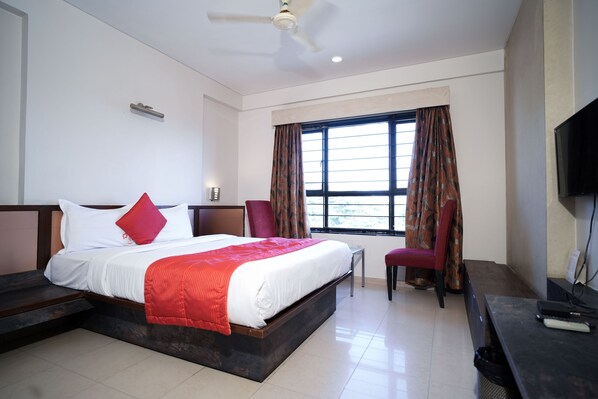 A hotel room with a large bed, a desk, a chair, a TV, and a window with curtains. - Cartier Biznotel (Bengaluru)