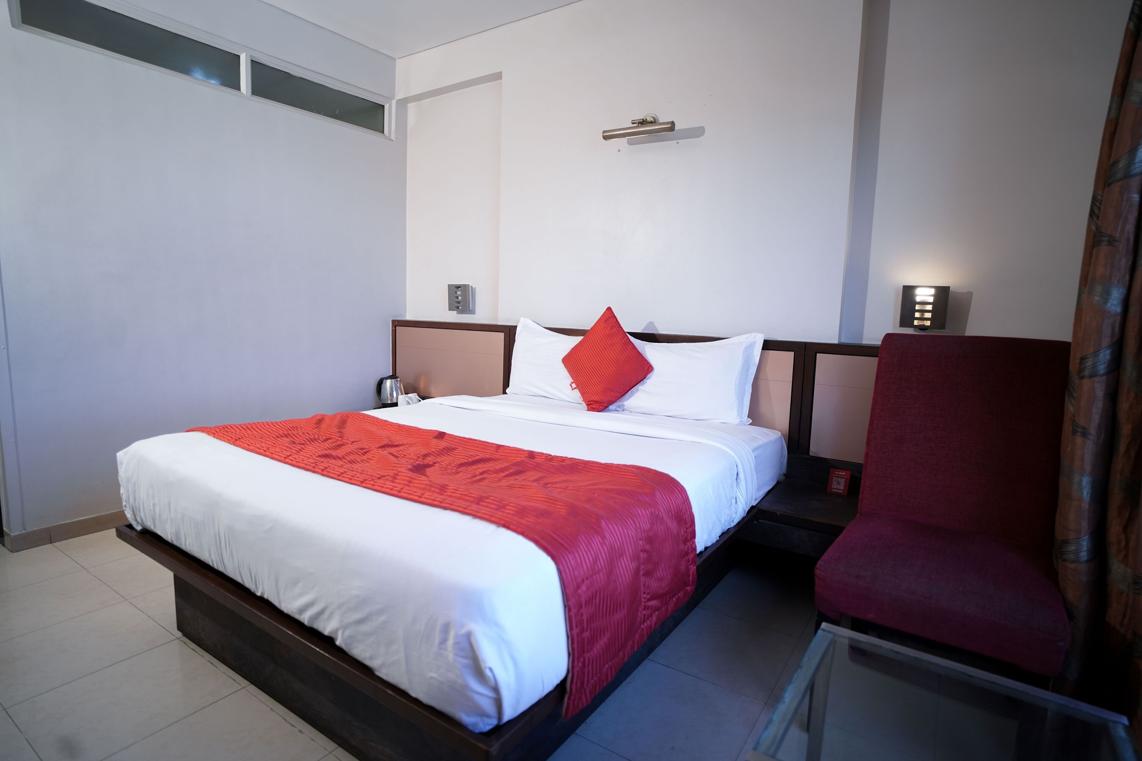 A hotel room with a large bed, a red chair, a bedside table, and a nightstand.