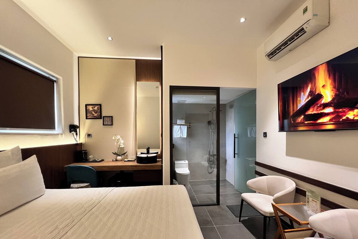 Une chambre d’hôtel moderne dotée d’un grand lit, d’un bureau, d’une chaise, d’un espace douche et d’un téléviseur diffusant une image de cheminée.