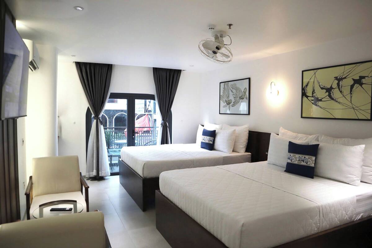 Une chambre d’hôtel avec deux lits, une chaise, une table, un ventilateur de plafond et un balcon avec des rideaux.