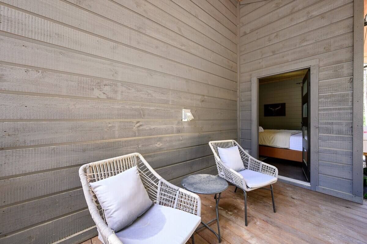 Een terras met rieten stoelen en een kleine ronde tafel, naast een houten muur en een deur naar een slaapkamer.