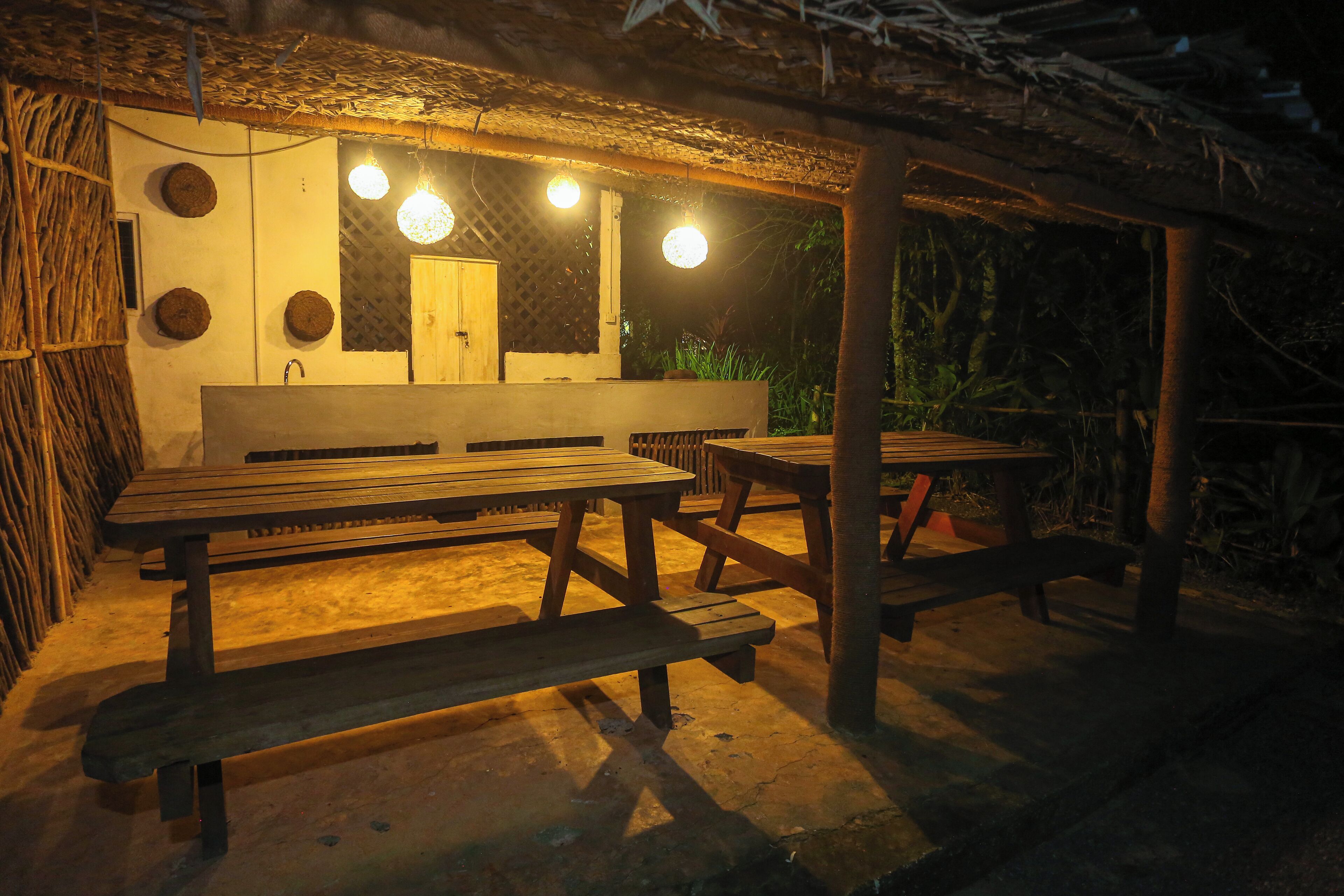 An outdoor seating area with wooden benches and tables, a thatched roof, and hanging lights.
