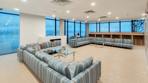 Un salon spacieux doté de grandes fenêtres, d’un canapé d’angle, d’une table basse en verre et d’un espace détente avec des bancs.