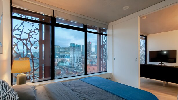 Une chambre moderne avec une grande fenêtre, un lit, une table de chevet avec une lampe et un téléviseur posé sur un meuble.