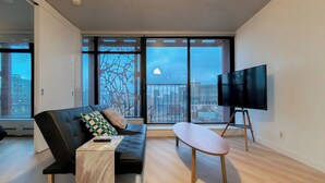 Un salon moderne avec un canapé en cuir noir, une table basse en bois et un téléviseur posé sur un meuble. De grandes fenêtres offrent une vue sur la ville.