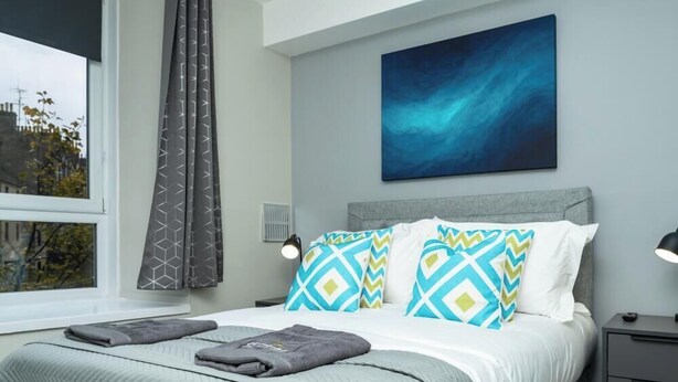 A modern bedroom with a bed, grey curtains, a blue abstract painting, and a city view. - City Road by Central Serviced Apartments (Dundee)