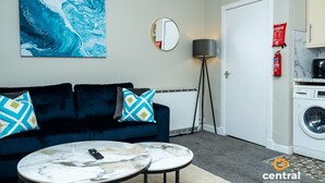 A modern living room with a dark blue sofa, two matching pillows, a round coffee table, a large abstract painting, a floor lamp, a washing machine, and a fire extinguisher. - City Road by Central Serviced Apartments (Dundee)