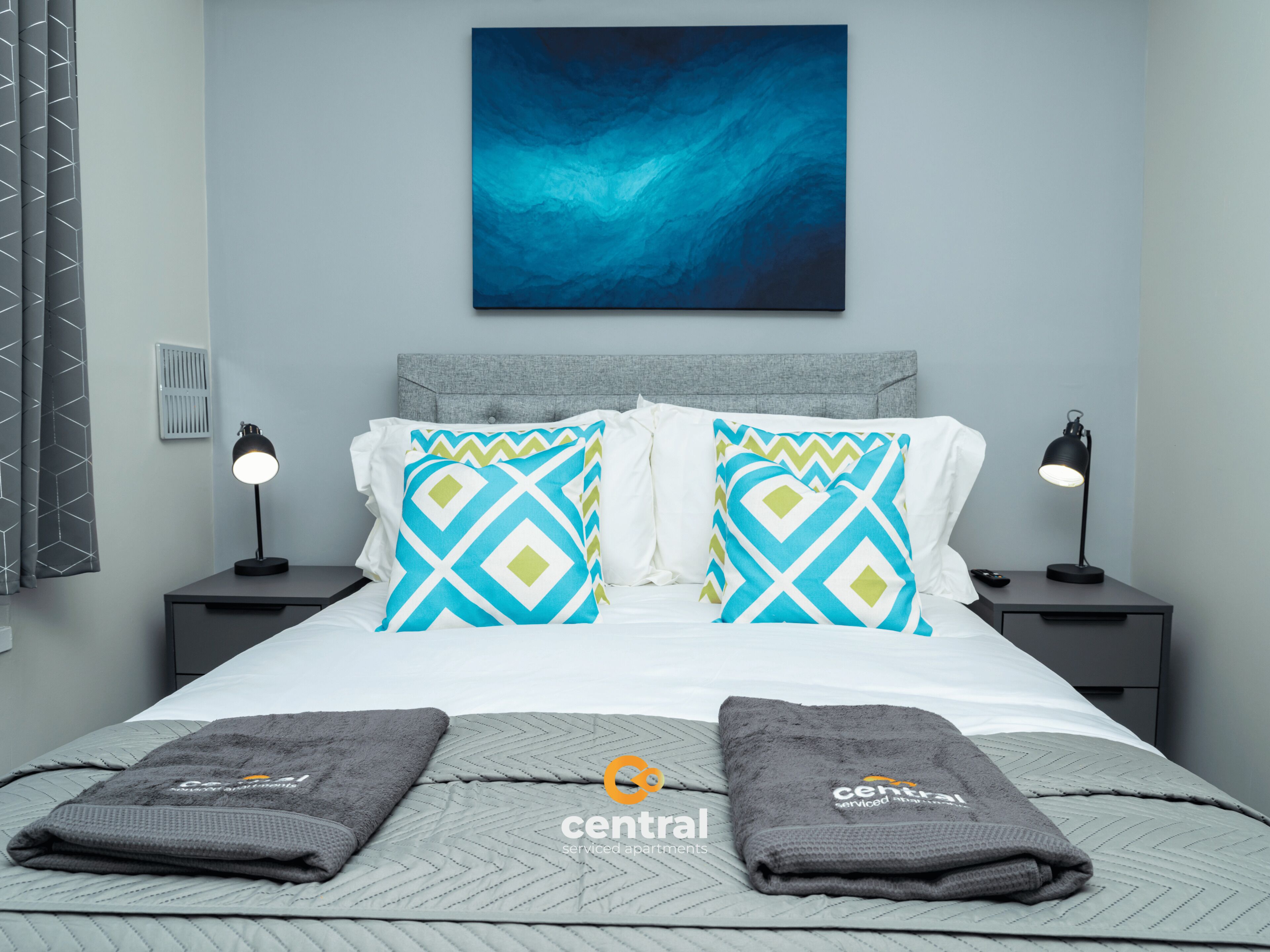 Un dormitorio moderno con cabecera gris, dos lámparas de noche, un cuadro abstracto azul y una colcha acolchada gris.