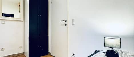 Una habitación con piso de madera, una puerta blanca, un armario oscuro, una mesita de noche con una lámpara y un espejo en la pared.