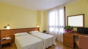 A hotel room with two single beds, a desk with a computer, a chair, a small bedside table with a lamp, and a potted plant. - hotel palas (Bruzolo)