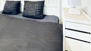 Una camera da letto con un letto bianco, due cuscini scuri, un comodino bianco con una lampada e un dreamcatcher appeso al muro.