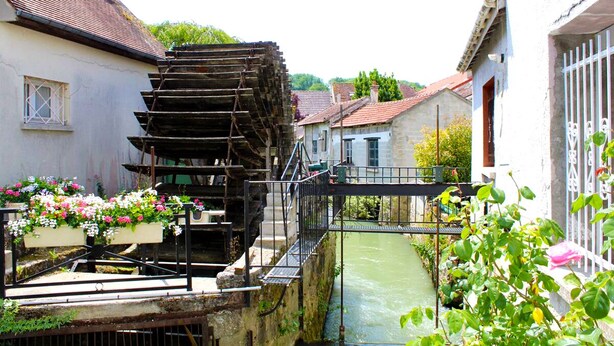 Un moulin à eau traditionnel, doté d’une structure en bois, entouré de bâtiments résidentiels et bordé d’un étroit canal.